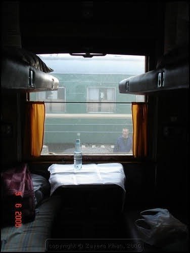 A train sleeper compartment with made-up bunks, a small table with a water bottle, and orange curtains.