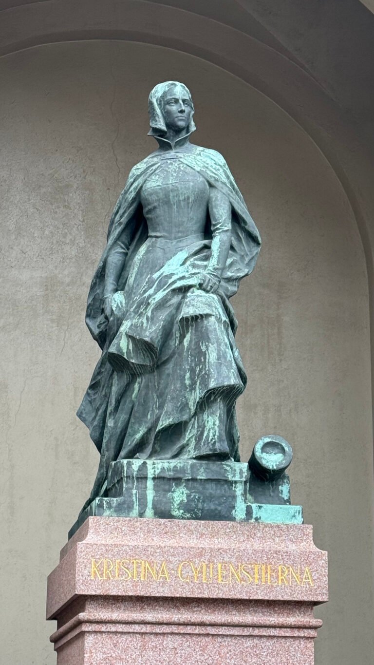 Bronze statue of Kristina Gyllenstierna at the Royal Palace in Stockholm. She wears a long dress, a cloak and a veil, and a cannon lies at her feet. The red granite base reads Kristina Gyllenstierna in gold letters.
