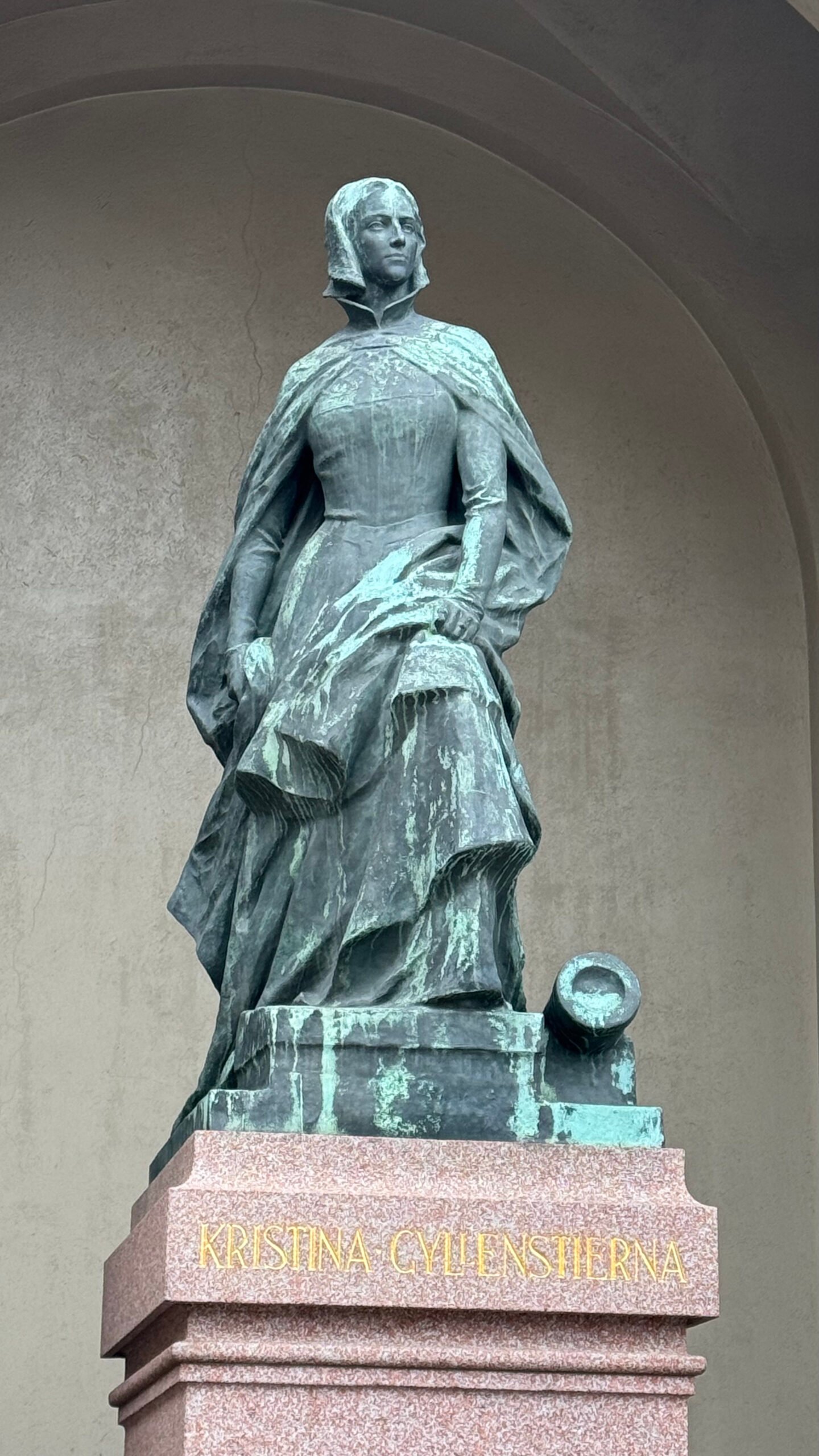 Bronze statue of Kristina Gyllenstierna at the Royal Palace in Stockholm. She wears a long dress, a cloak and a veil, and a cannon lies at her feet. The red granite base reads Kristina Gyllenstierna in gold letters.