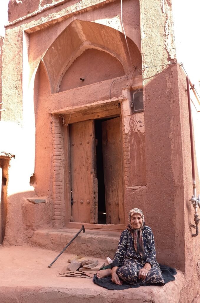 Older woman in a flower scarf sits by a wooden door in Abyaneh
