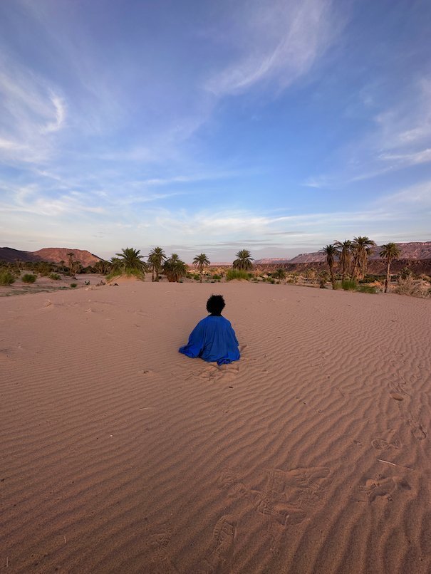 A man in a blue robe sits on a sand dune and looks at palm trees.