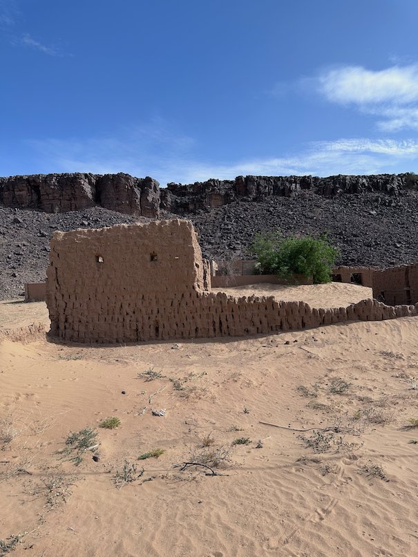An old mud brick wall stands in sand below a dark rock ridge.