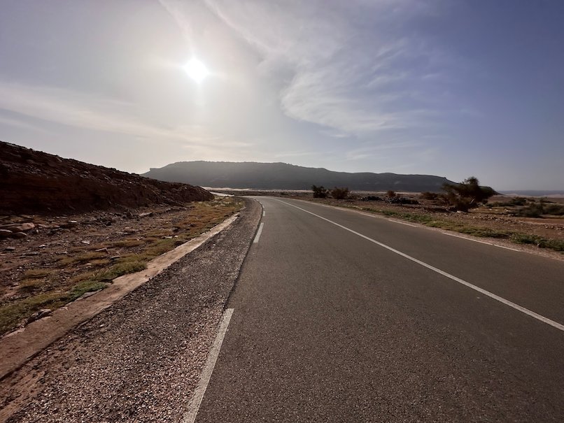 The sun shines through haze over a road and a flat mountain.