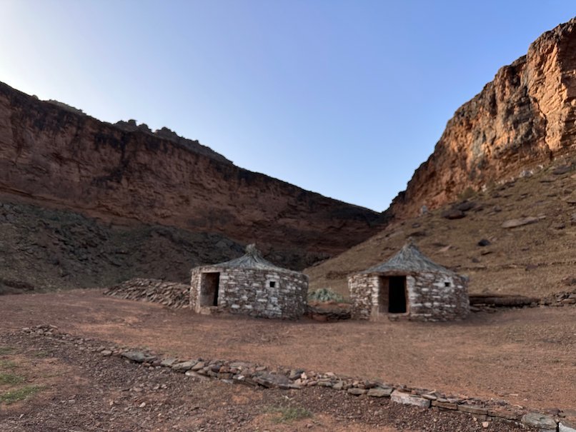 Two small stone huts stand at the foot of red rock cliffs.