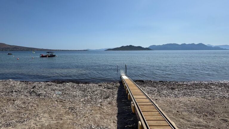 A narrow wooden jetty leads from a pebble beach into the calm Saronic Gulf on Aegina, with islands and mountains on the horizon