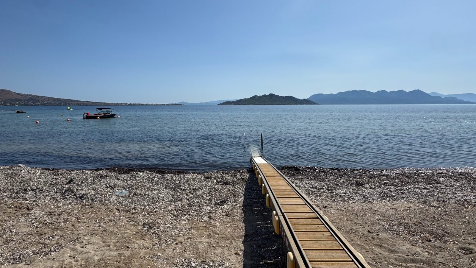 A narrow wooden jetty leads from a pebble beach into the calm Saronic Gulf on Aegina, with islands and mountains on the horizon