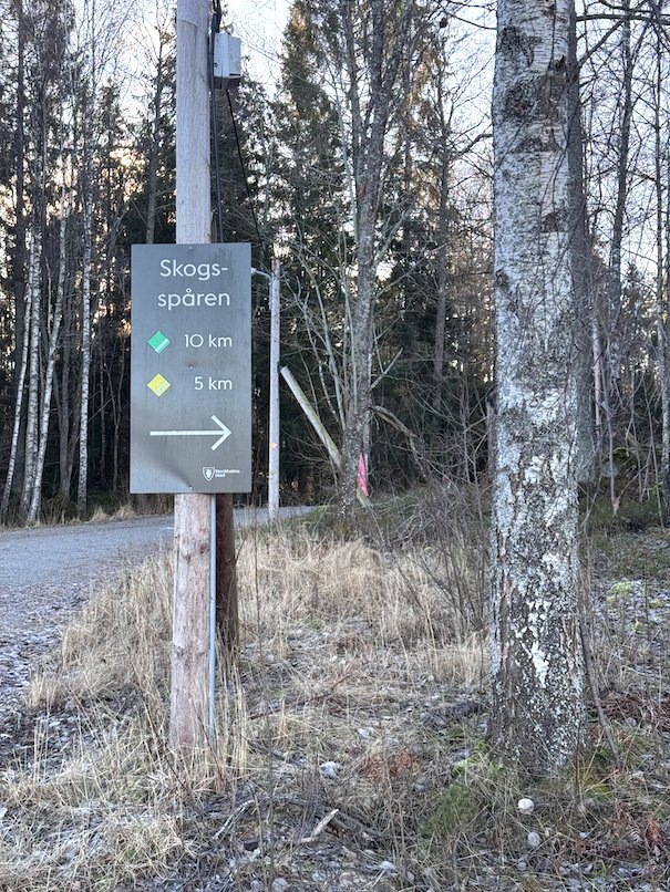 Trail sign Skogsspåren with arrows for the 10 km and 5 km routes, on a post by a gravel road in Ågesta nature reserve