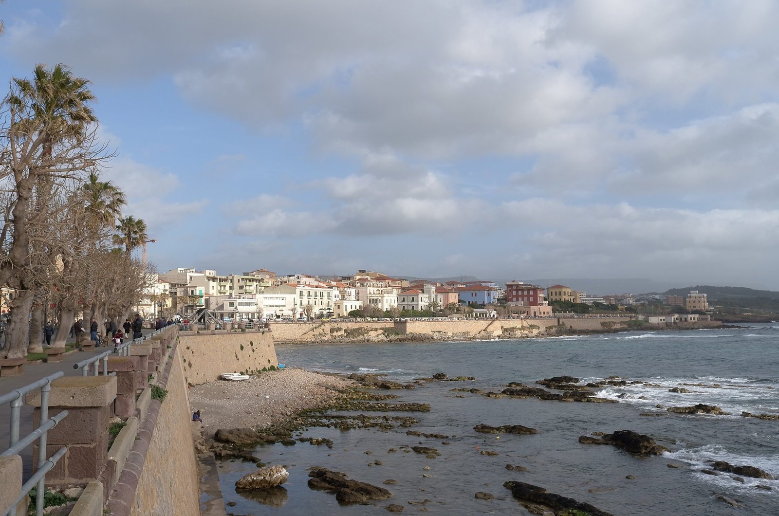 A promenade with palm trees runs along the rocky coast toward Alghero town.