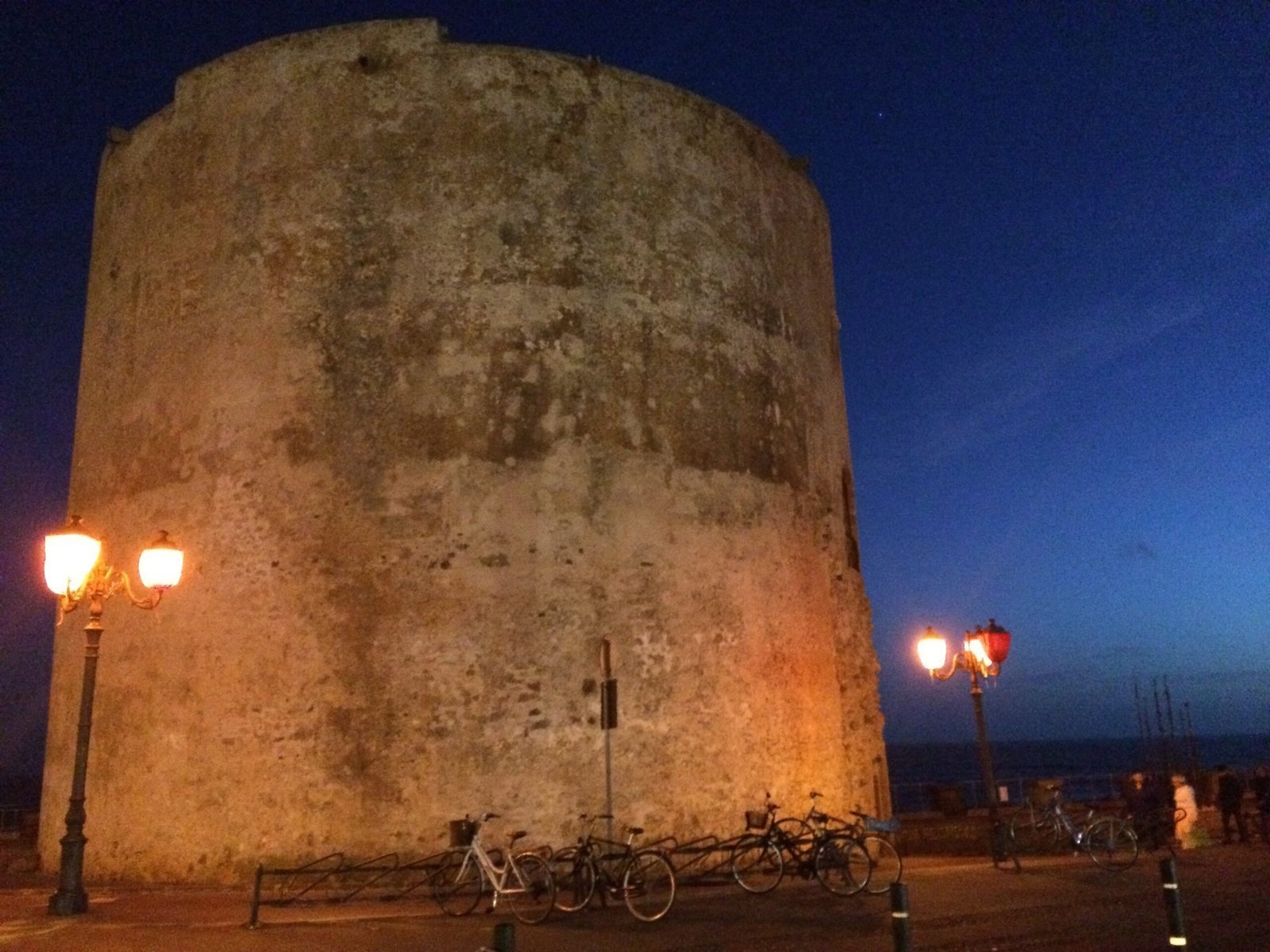 A round stone tower stands under a blue evening sky on the promenade in Alghero.