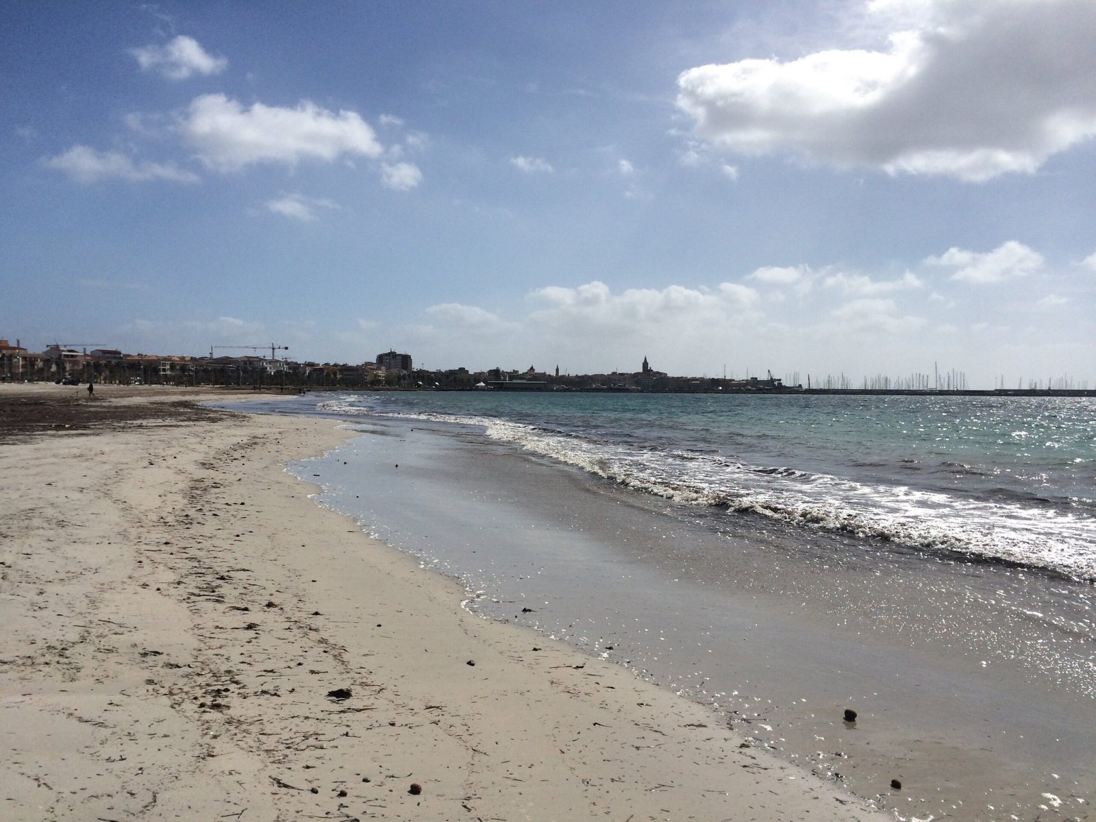 A sandy beach with small waves curves toward the town of Alghero.