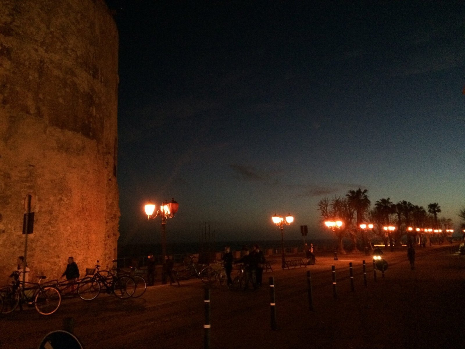 A stone tower and street lamps stand on the seafront promenade in Alghero at dusk.