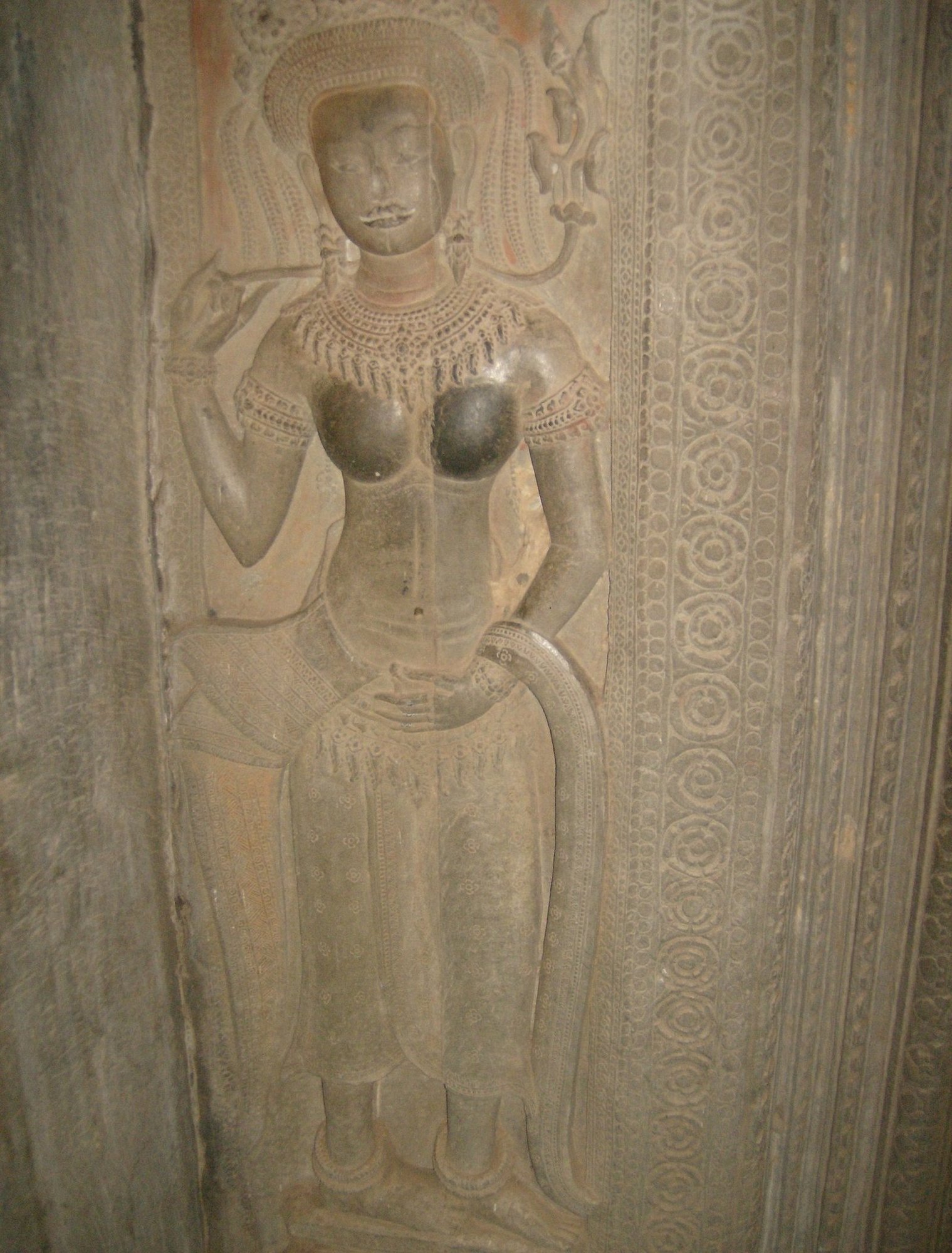 A carved female figure with a tall headdress stands on a wall at Angkor Wat.