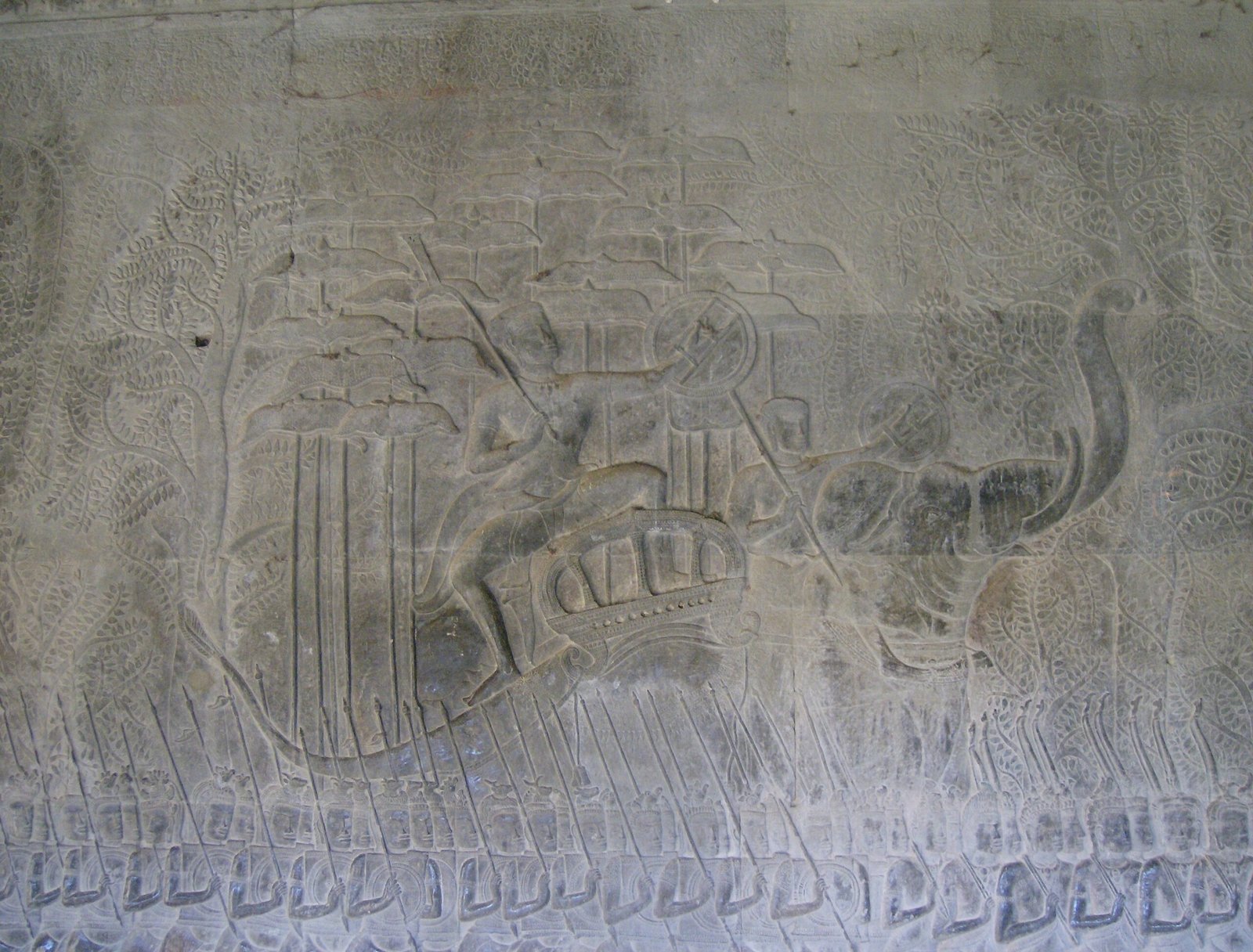 A carved stone relief shows warriors and a chariot on a gallery wall at Angkor Wat.