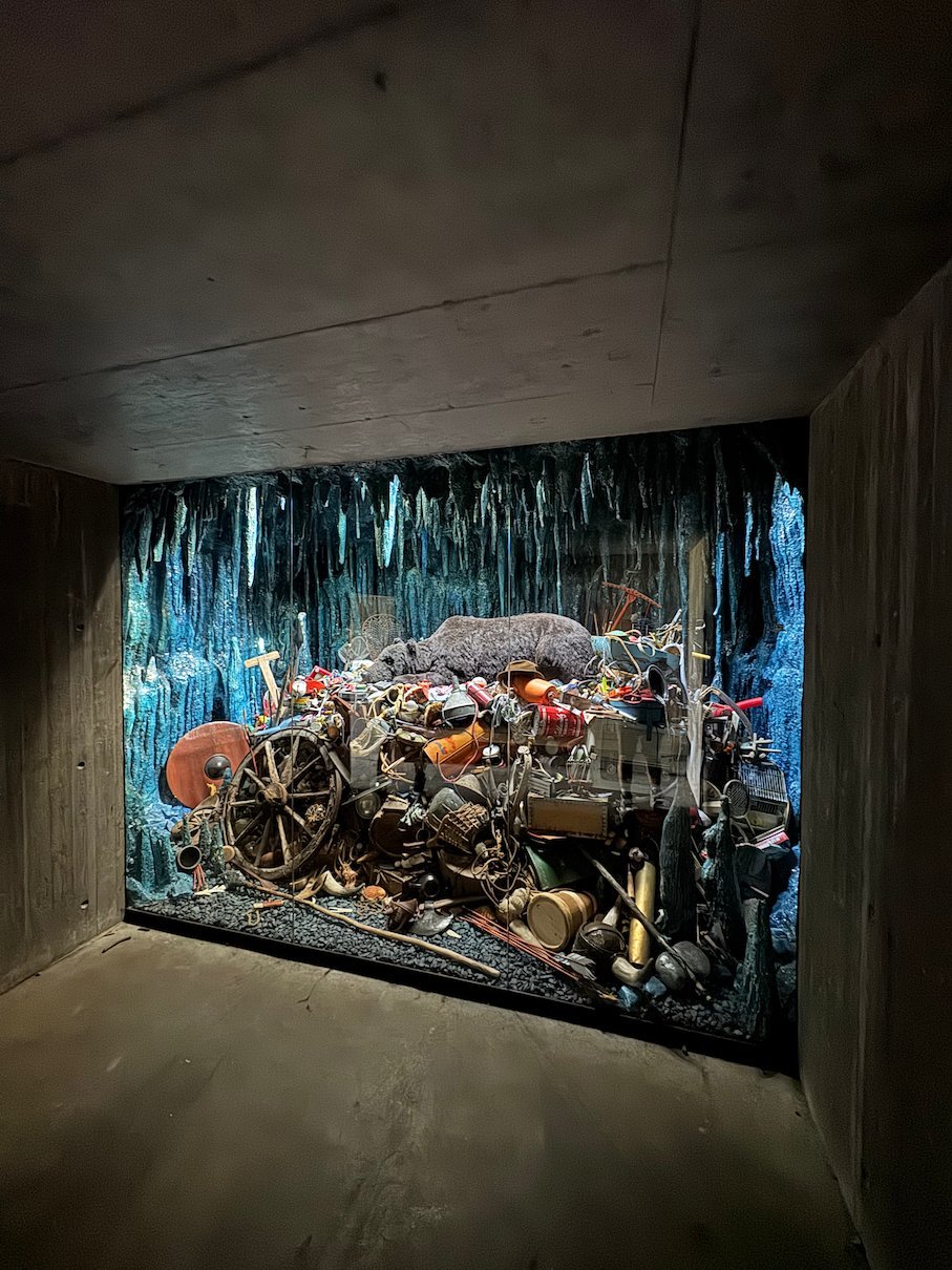 Art installation with many objects behind glass in a small dark room by the road