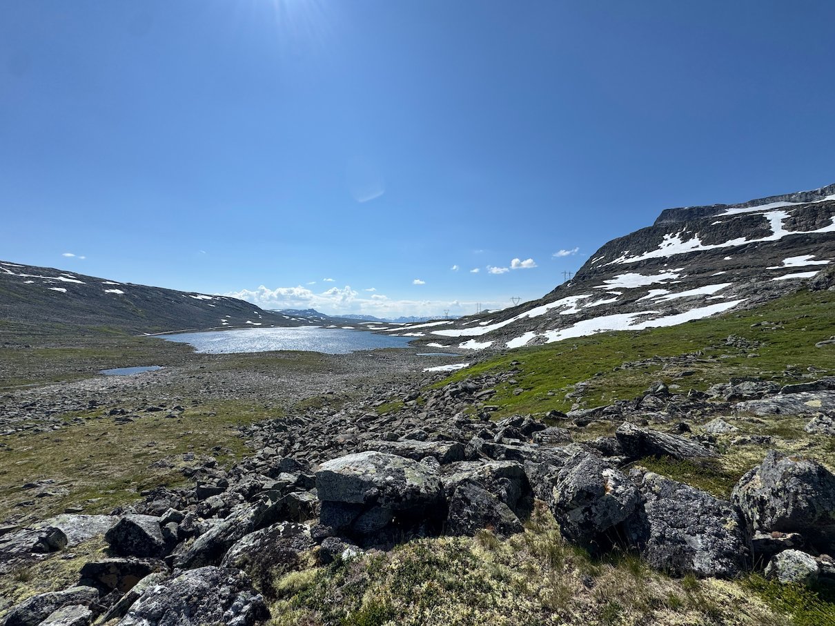 Plateau with rocks, a long lake and snowy hills under a blue sky