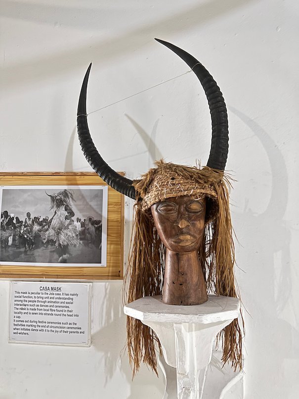 A mask with long horns and a fibre wig stands on a stand in the Kachikally museum, Bakau.