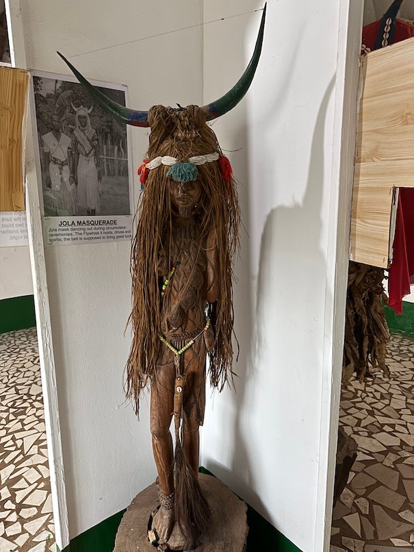 A horned masquerade figure in fibre dress stands in the museum at Kachikally, Bakau.
