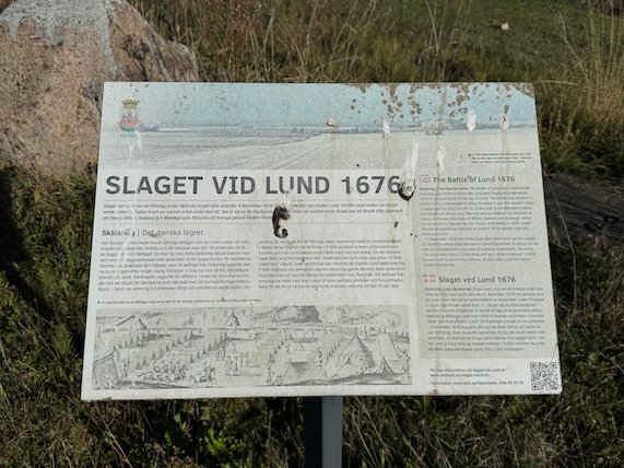 Weathered information sign about the Battle of Lund 1676 beside a large stone in a field