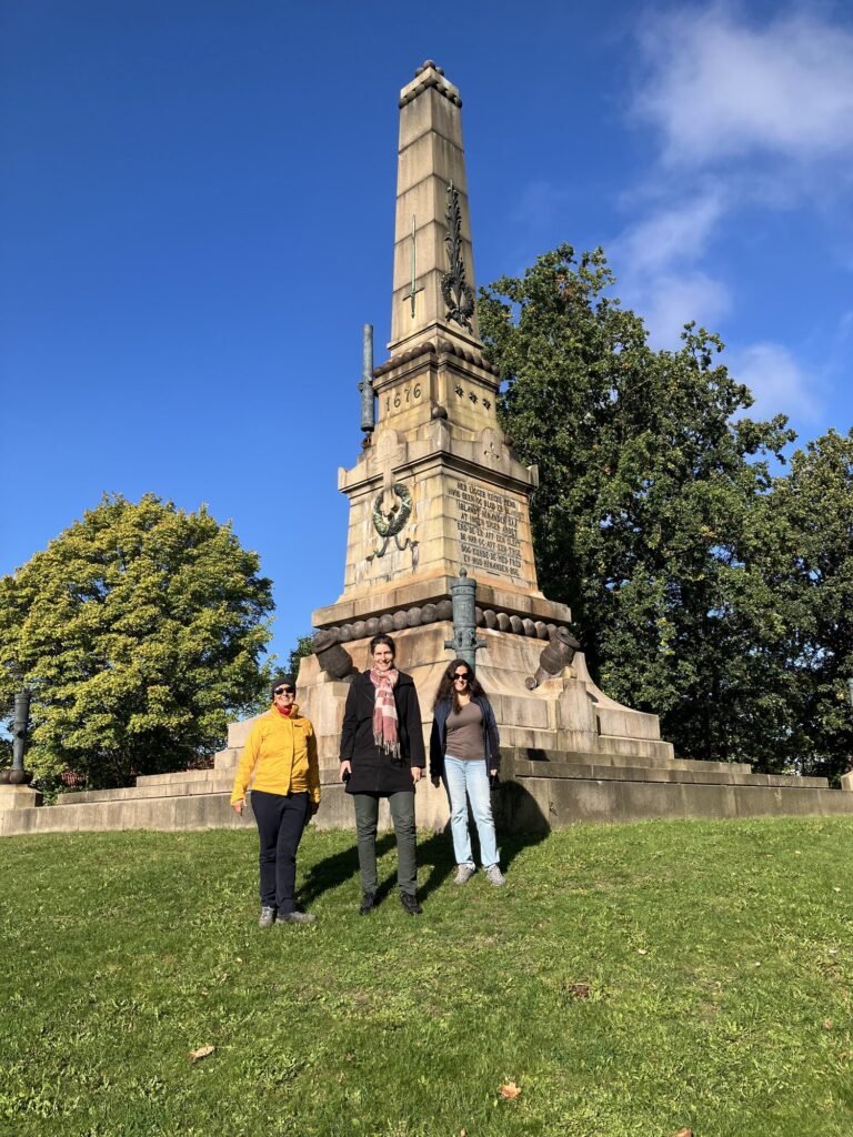Three people stand in front of the Battle of Lund obelisk on the grass hill Lerbäckshög in Lund