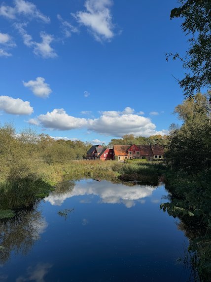 Calm stream that reflects white clouds, with red brick farm buildings behind the trees