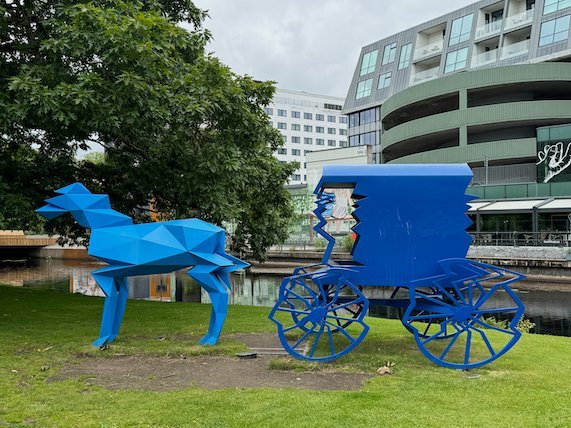 Blue geometric sculpture of a horse and carriage on a lawn by the river
