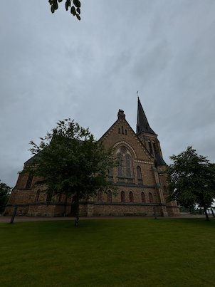 Brick church with a tall spire behind trees and a lawn in Borås