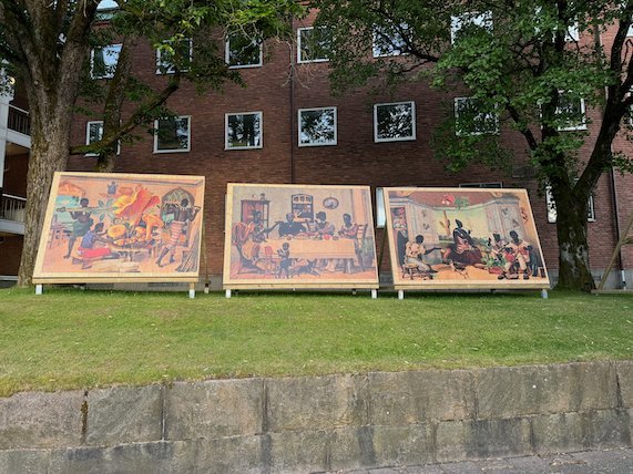 Three large painted panels by Gê Viana on the grass in front of a brick building