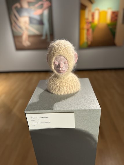 Head sculpture in a knitted cream hood on a white plinth