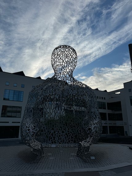 Large sculpture of a seated figure made of open metal mesh, in a square in Borås under a cloudy sky