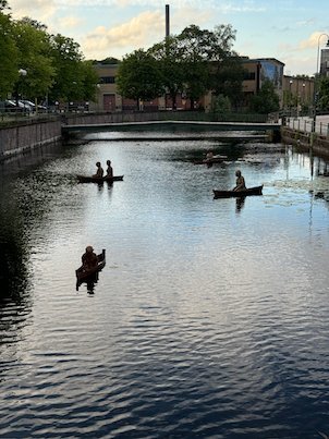 Figures in small boats on the calm Viskan river in Borås, part of an art installation