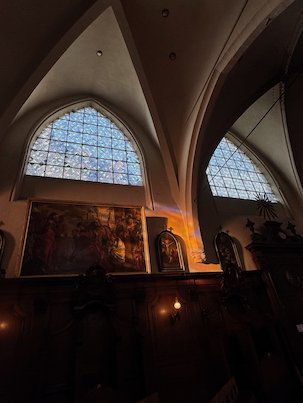 A pale window lights a vaulted wall above old paintings in a church in Brussels.