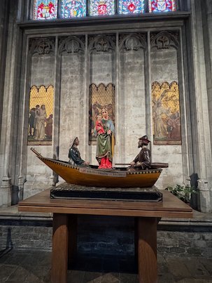 A wooden boat with figures stands in front of painted panels in the Sablon church.