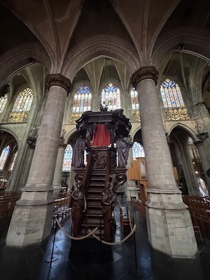 A carved wooden pulpit stands between stone pillars in the Sablon church in Brussels.