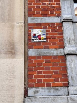 A small colorful tile hangs on a red brick wall between stone columns in Brussels.
