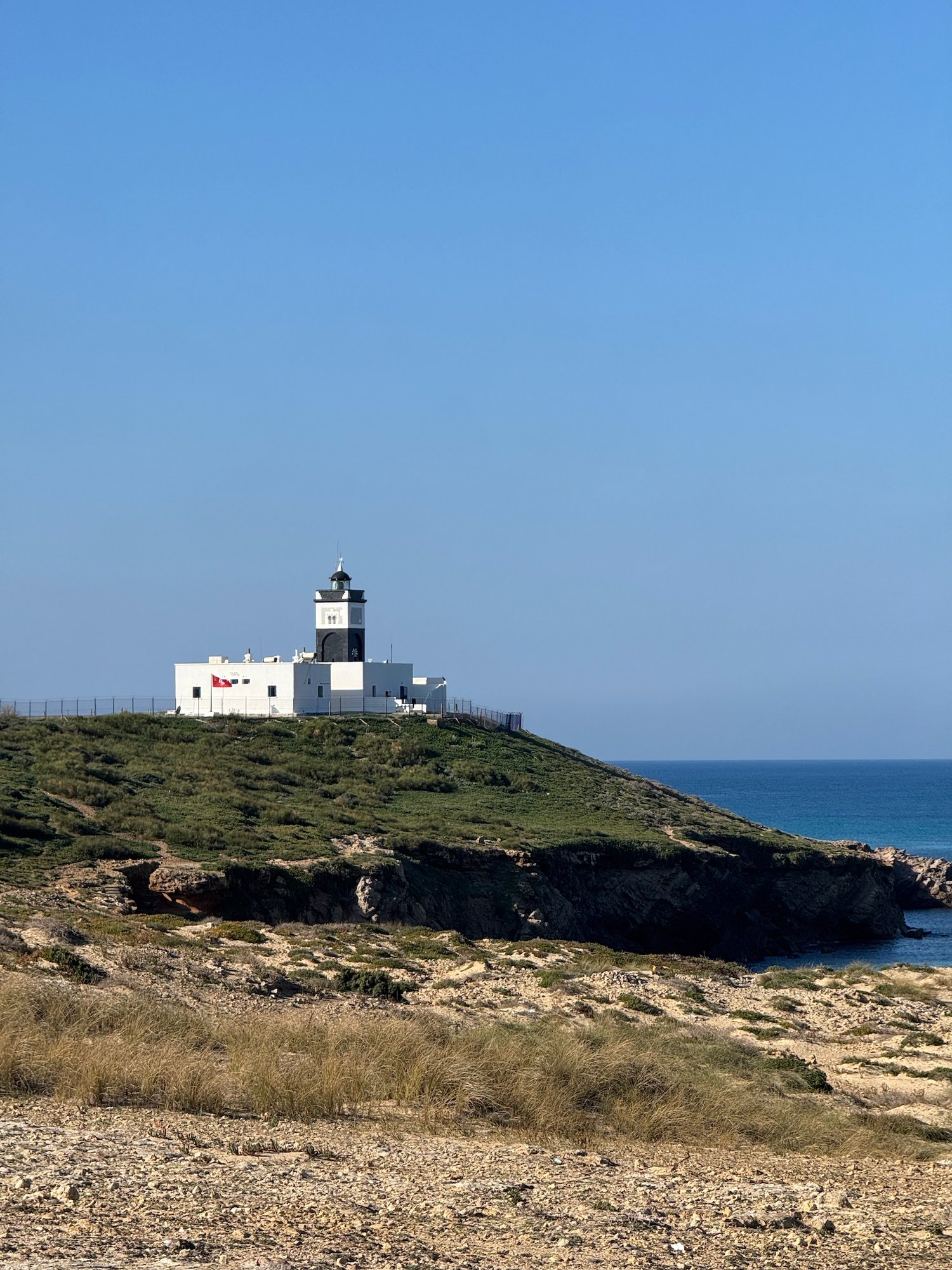 The black and white lighthouse tower and keeper house stand on a green cliff at Cap Angela, Tunisia.