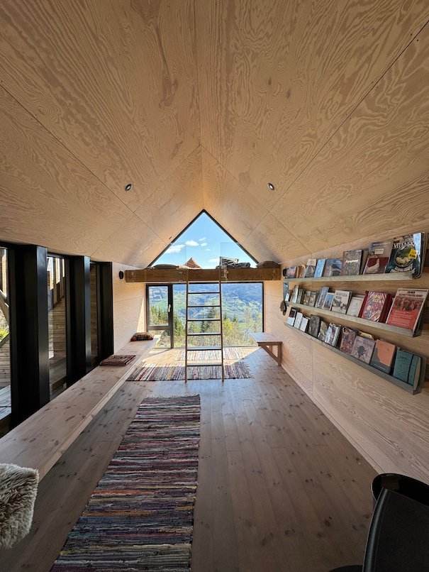 Inside Dagsturhytta: light wooden walls, benches, a long rug and a large window toward the mountains