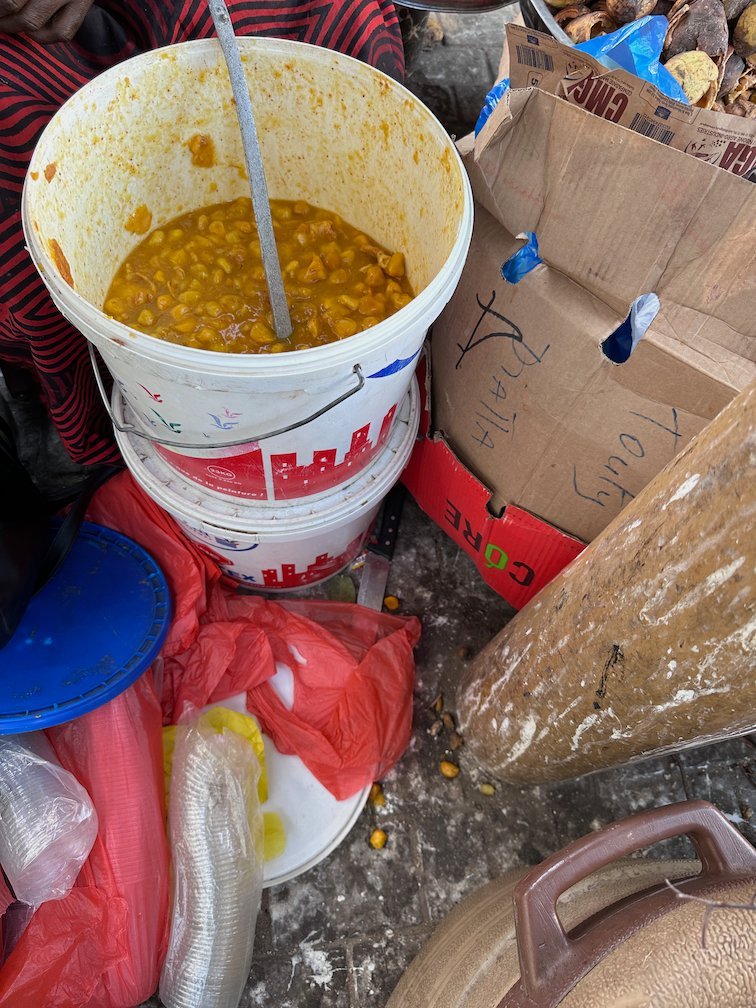 A bucket of cooked beans stands among bags and boxes at a market in Dakar.