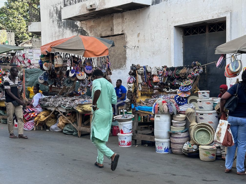 A man in green walks past stalls with baskets and pots on a street in Dakar.