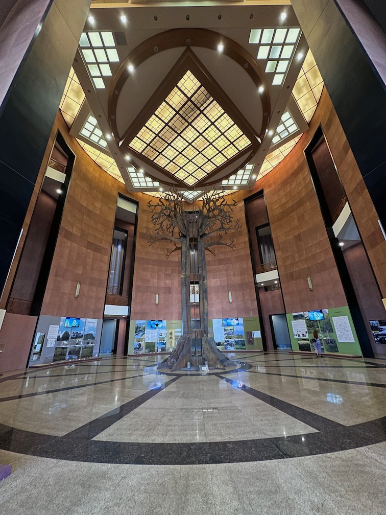 A tall round hall with a glass ceiling and a tree sculpture in a Dakar museum.