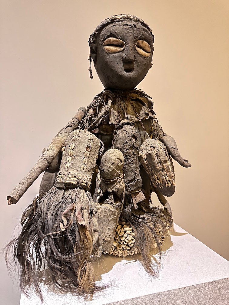 A figure with a dark face, shells and fibers stands in a Dakar museum.
