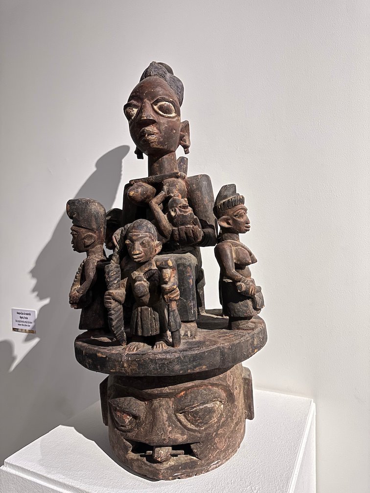 A wooden mask with many small carved figures on top stands in a Dakar museum.