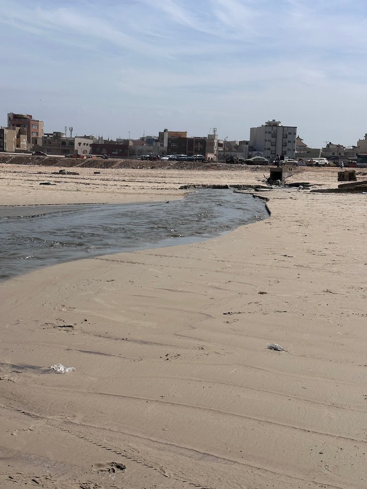 Dark water flows in a channel across the beach toward the sea in Dakar.