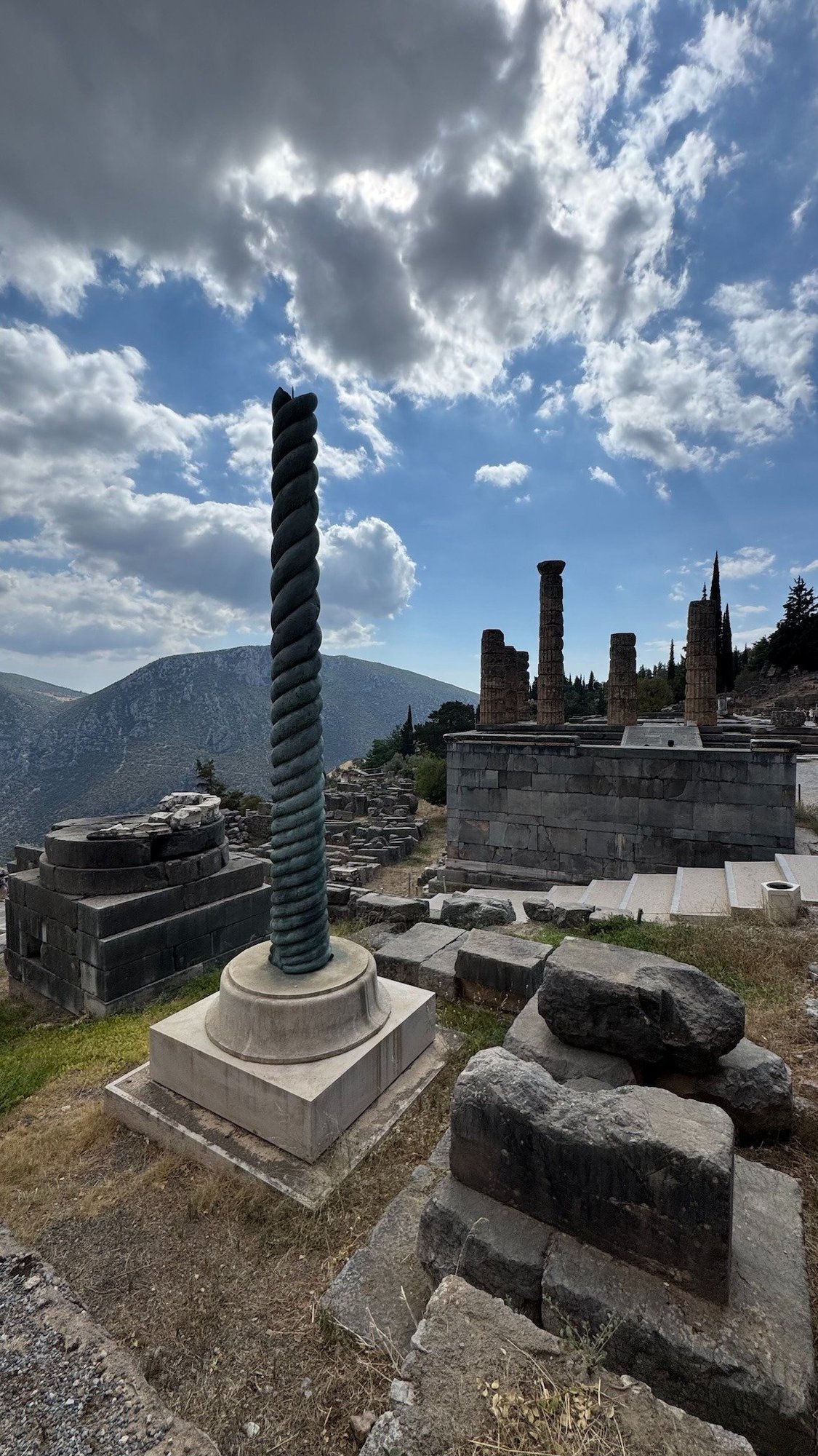 Spiral bronze-colored column in the Sanctuary of Apollo at Delphi, with ruins and mountains around it