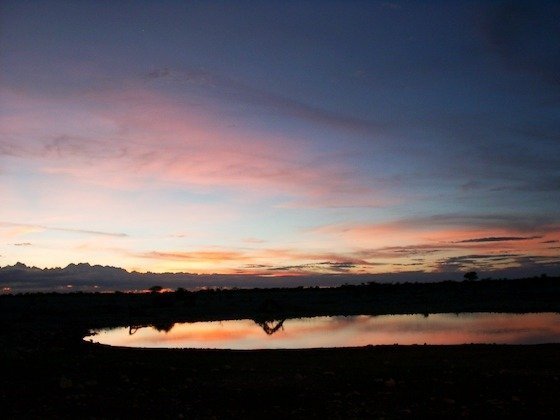 Pink and orange clouds shine over a dark waterhole at sunset in Etosha, Namibia.