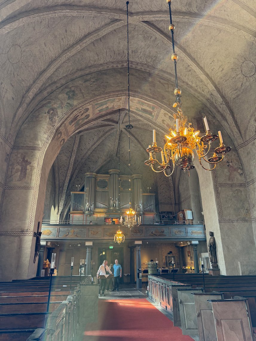 Interior of Gamla Uppsala church, with white vaults, a brass chandelier and the organ at the west end