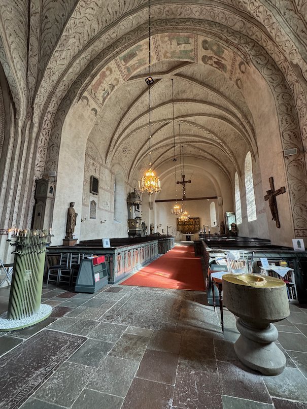 Nave of Gamla Uppsala church with a stone baptismal font, pews and a red carpet to the altar