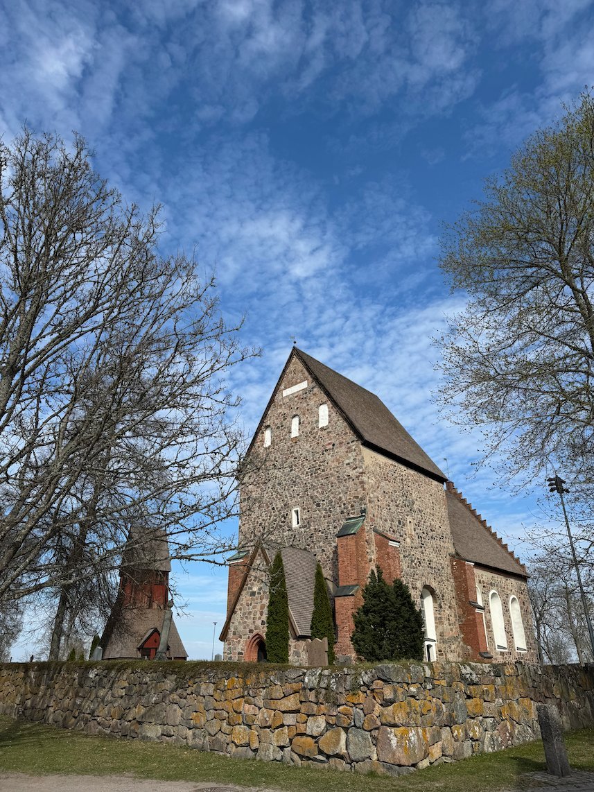 Gamla Uppsala church with its stone walls and red brick gable, behind a dry stone wall under a blue sky