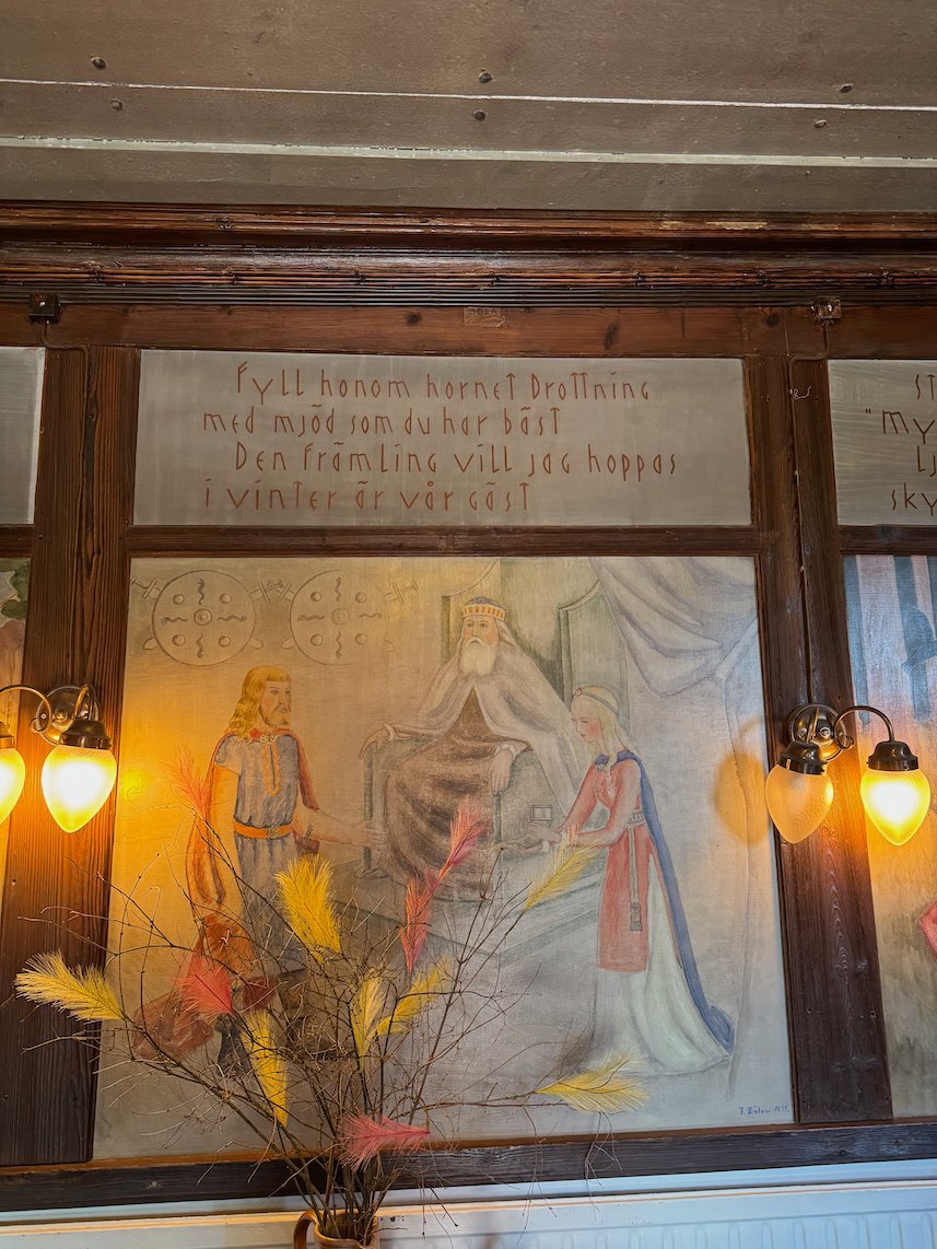Painted wall panel at Odinsborg with Norse gods and old Swedish verse above, lit by wall lamps