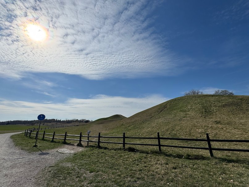 Grass-covered royal mound with a wooden fence and a gravel path, sun in a blue sky