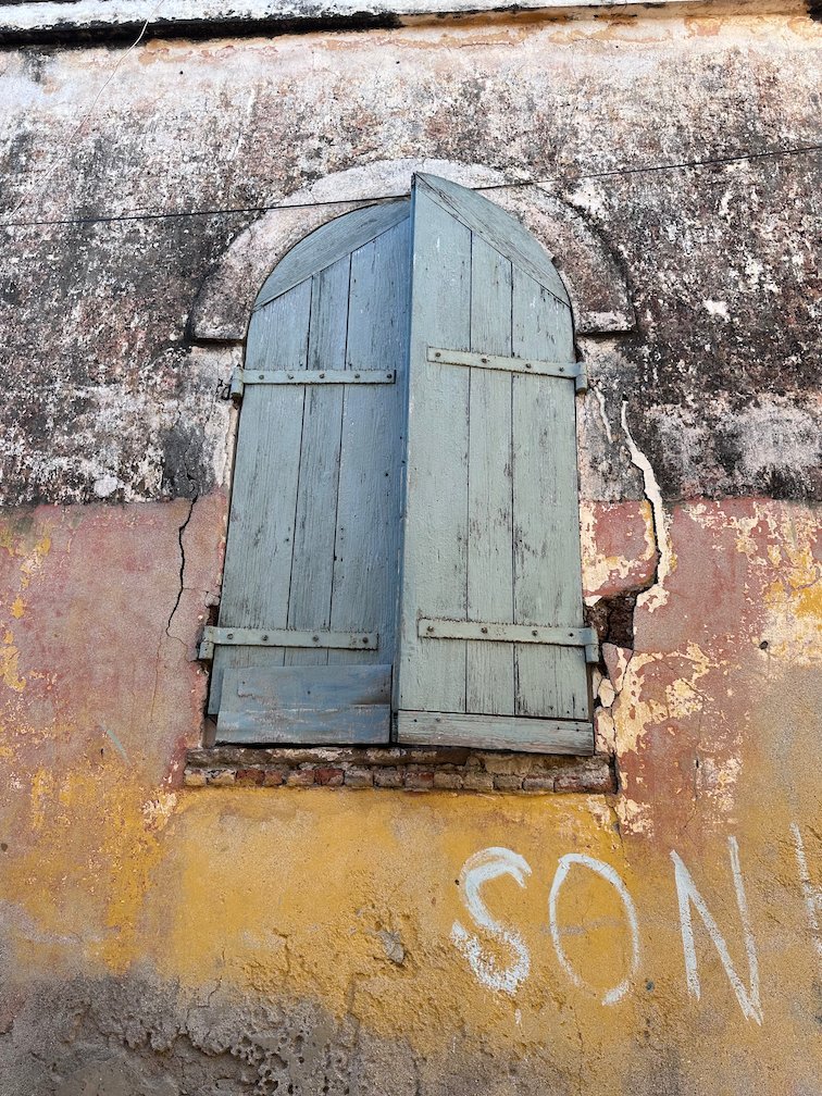 A blue wooden shutter covers a window in an old wall on Goree Island.